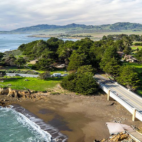 A scenic coastal landscape featuring a bridge, lush greenery, and distant mountains under a cloudy sky.