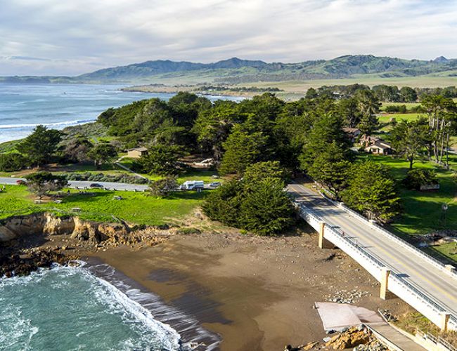 A scenic coastal landscape featuring a bridge, lush greenery, and distant mountains under a cloudy sky.