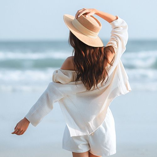 A person in a hat and light clothing stands on a beach, facing the ocean, with waves in the background.