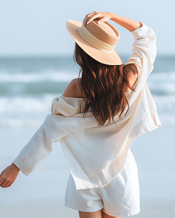A person in a hat and light clothing stands on a beach, facing the ocean, with waves in the background.