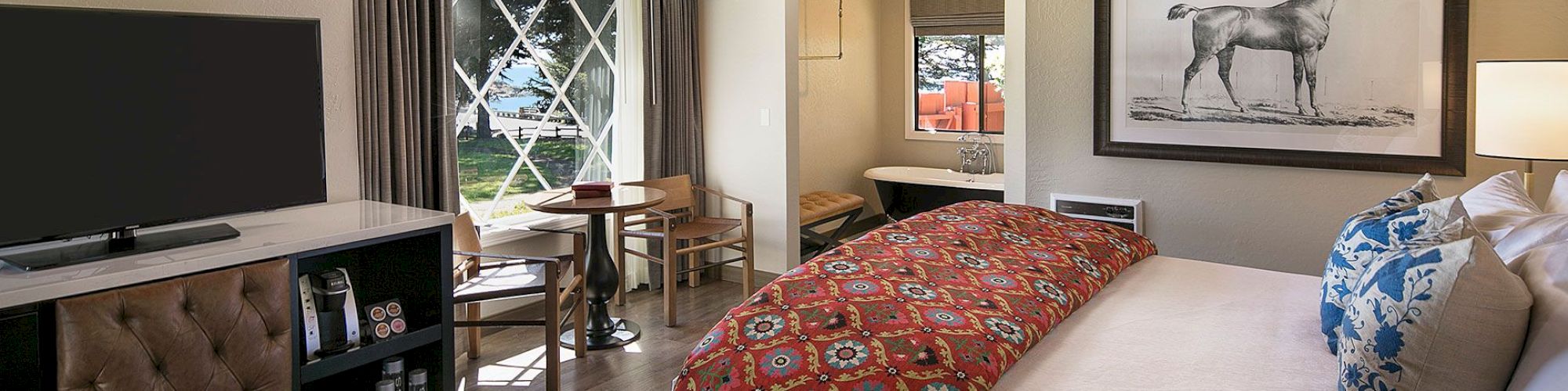 A hotel room with a large bed, patterned red comforter, flat-screen TV, dresser, seating area, and a framed horse drawing on the wall.