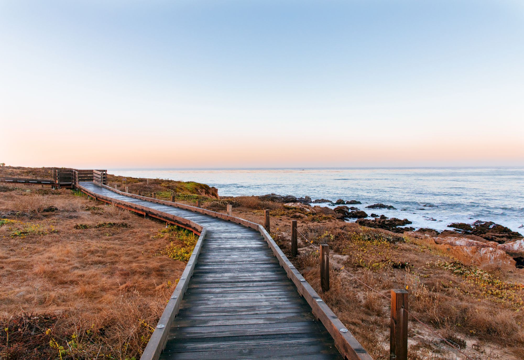 A wooden boardwalk winds along a rocky coastline at sunset, with dry grass, calm seas, and a distant horizon.