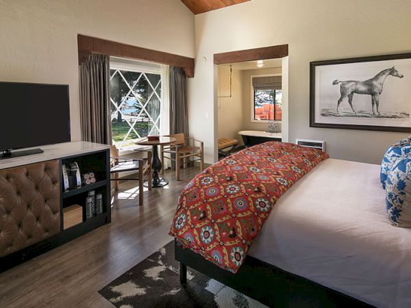 A cozy hotel room with a bed, a patterned red duvet, a TV stand, small desk and chairs, and a framed horse print on the wall, warm lighting.