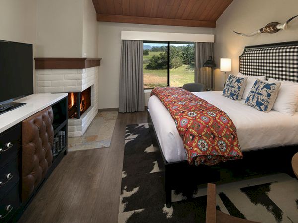 A cozy hotel room with a king bed, patterned throw, fireplace, TV cabinet, sliding glass door to a garden view, and a deer antler wall art.