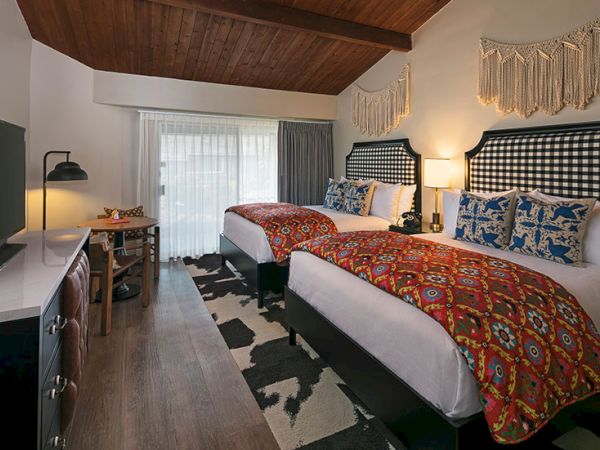 Two double beds in a cozy, rustic hotel room with patterned red bedding, woven headboards, a desk, TV, and a windowed door to a balcony.