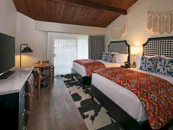 Two tidy hotel rooms with two queen beds, patterned red bedspreads, blue pillows, a wooden ceiling, desk, TV, and a sliding glass door to a balcony, bright and cozy.