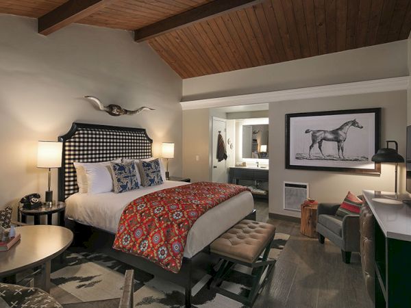 A stylish hotel room with a king bed, plaid headboard, red patterned throw, two bedside lamps, a bench at the foot, and a seating area with a framed horse art in the corner.