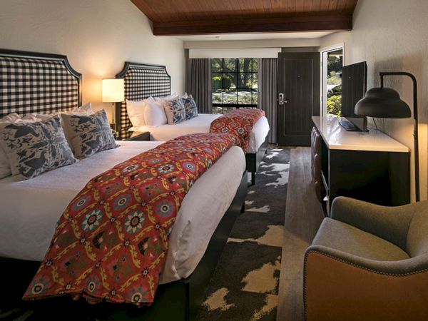 Two queen beds with patterned headboards, red floral runners, desk, lamp, armchair, and a doorway to a garden view&mdash;cozy hotel room.