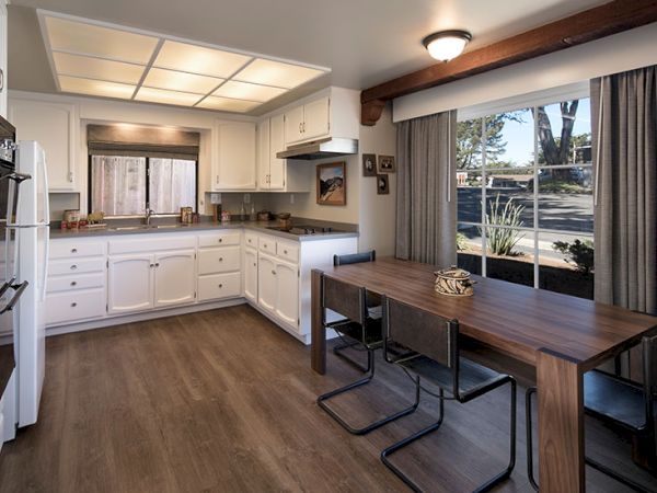 A bright L-shaped kitchen with white cabinets, a wooden dining table, dark flooring, large window with curtains, and warm ceiling lighting.