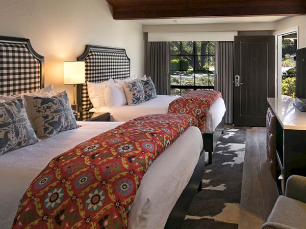 Three neatly made hotel beds with red patterned runners, nightstand lamps, and a view into a doorway and curtains at the far end.