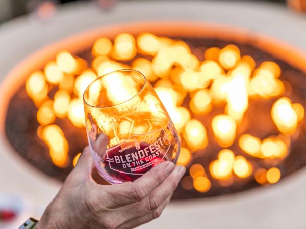 A hand holds a glass of whiskey with ice, blurred flames glow in a fire pit background, label reads &ldquo;BLENDERS&rdquo; on the glass, warm ambiance.