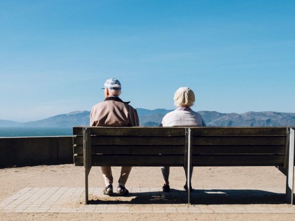 Two elderly people sit on a bench by the waterfront, facing the sea and distant mountains on a sunny day.