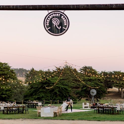 A cozy outdoor wedding or event setup with string lights, tables, chairs, and greenery under a wooden gate at dusk.