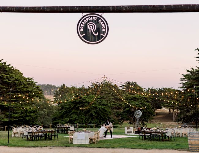A cozy outdoor wedding or event setup with string lights, tables, chairs, and greenery under a wooden gate at dusk.