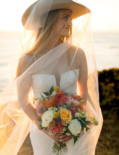 A bride in a white gown and veil holds a colorful bouquet at sunset by the coast, smiling softly.