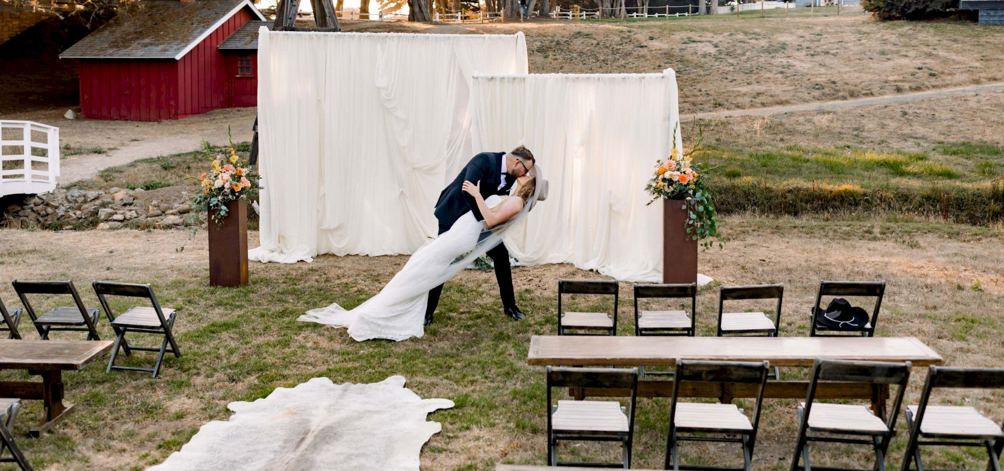 A couple shares a kiss during an outdoor wedding ceremony setup, with chairs, a white backdrop, and a windmill in a sunny field.