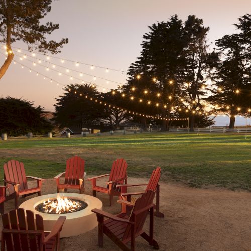 A cozy outdoor gathering around a lit fire pit with string lights and red Adirondack chairs in a park at dusk, inviting warmth and conversation.
