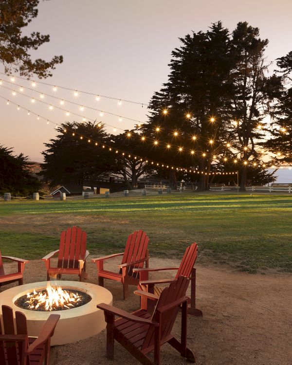 A cozy outdoor gathering around a lit fire pit with string lights and red Adirondack chairs in a park at dusk, inviting warmth and conversation.
