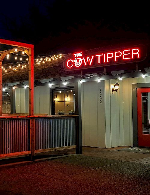 A nighttime scene of The Cow Tipper restaurant with string lights, an outdoor seating deck, and a red lit sign at the entrance.