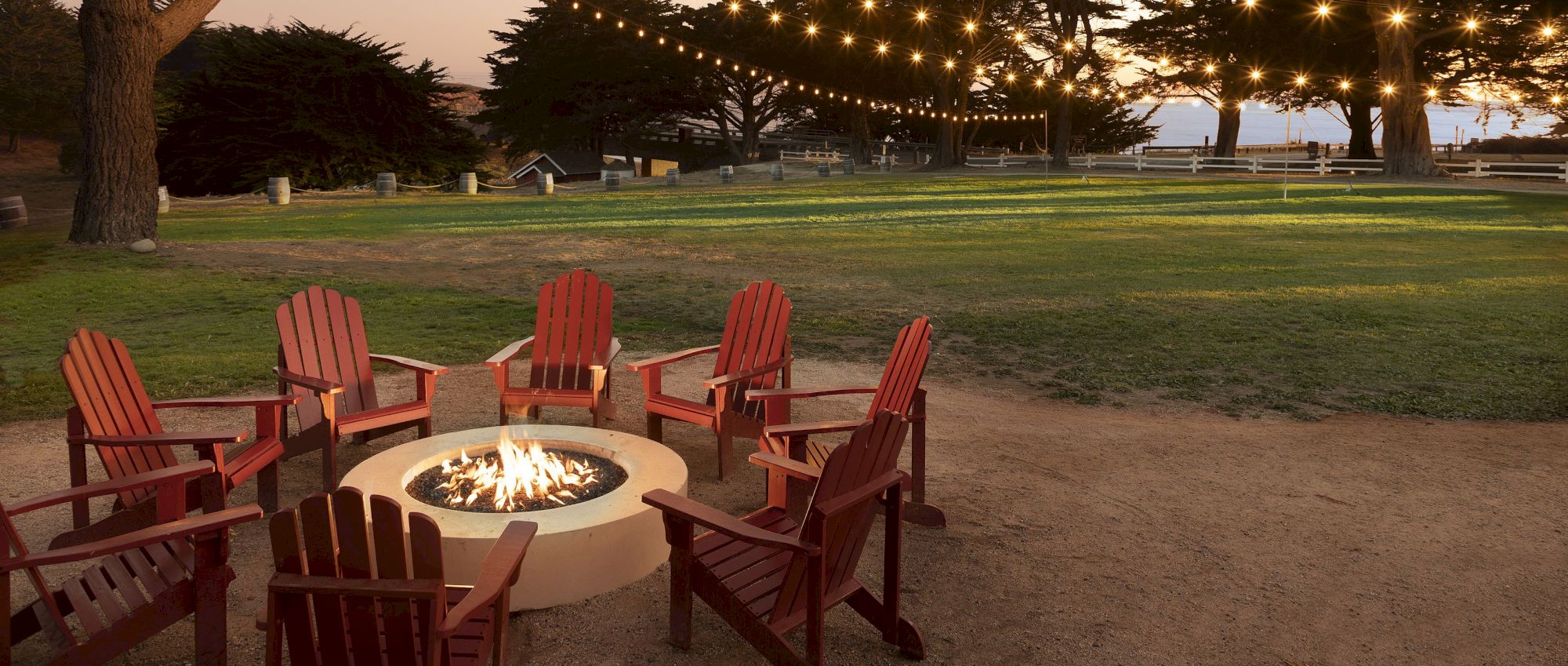 A cozy outdoor setup with a circular fire pit and red Adirondack chairs arranged around it, string lights overhead in a park at dusk.