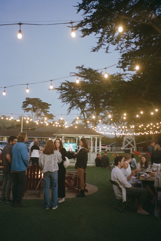 A lively outdoor evening gathering under string lights, people chatting and dining at tables on a lawn with trees and a clear sky.