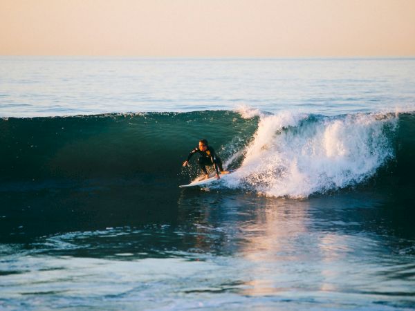 A surfer rides a turquoise wave at dusk, carving along the lip as water glints orange on the horizon, calm sea stretching beyond.