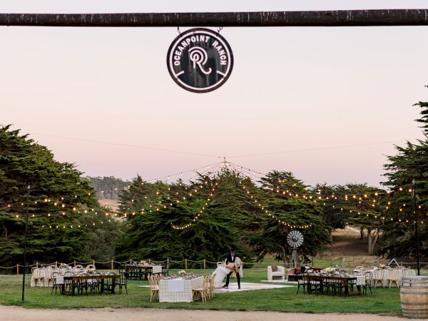 An outdoor wedding setup with tables, string lights, and greenery under a circular sign hanging above; a couple in the center and a scenic park backdrop.