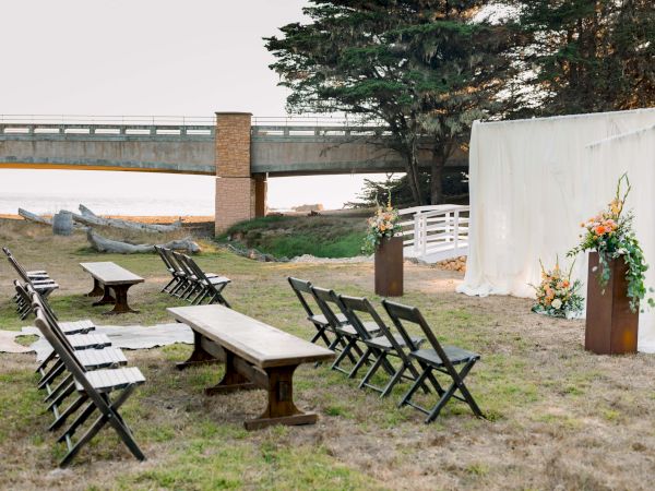 An outdoor wedding setup with empty black chairs, a wooden table, a white fabric backdrop, flower arrangements, and a coastal bridge in the background.