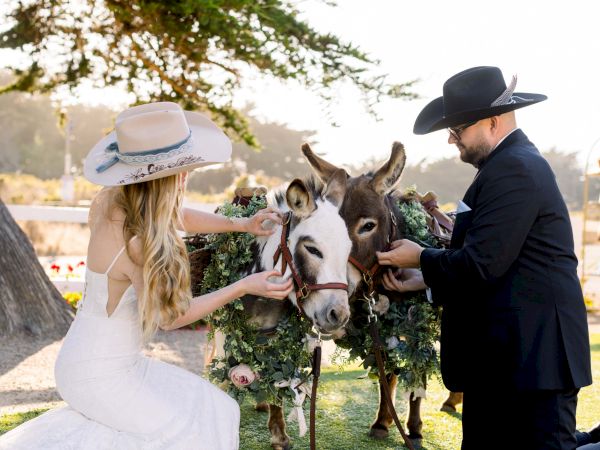 Two people in wedding attire posing with a decorated donkey outdoors, the bride and groom gently holding its head near a floral garland.