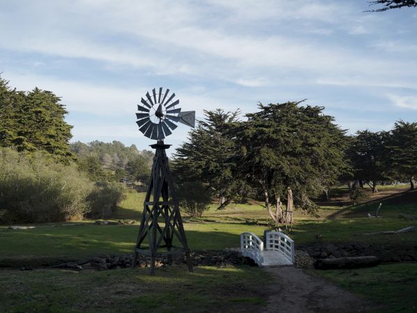 A wooden windmill beside a small white gate on a grassy field with trees and a blue sky in the background.