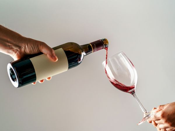 Two hands pour red wine from a bottle into a tilted glass against a light background.