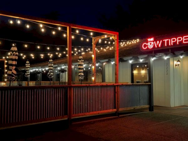 A lit outdoor patio of a restaurant or bar at night, string lights overhead, wooden railing, and a neon sign reading &ldquo;The Cow Tipper&rdquo; on the building.
