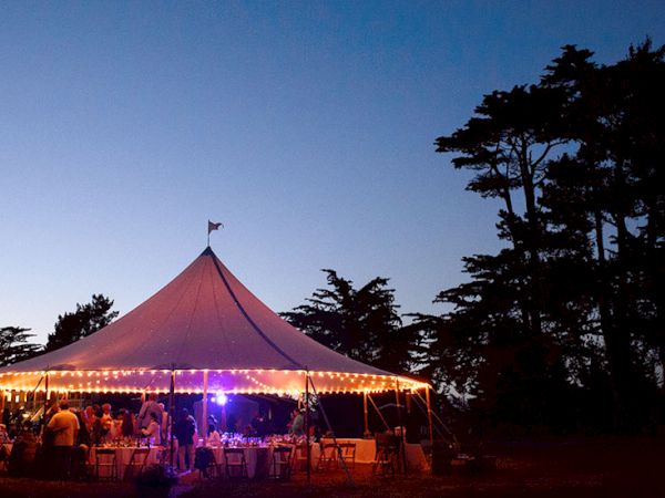 A large white event tent with string lights, people seated around tables, evening sky, tall trees in the background, festive outdoor gathering.