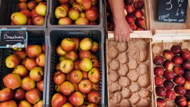 A street market scene with crates of apples and pears on the left, and eggs and peaches in paper trays on the right.