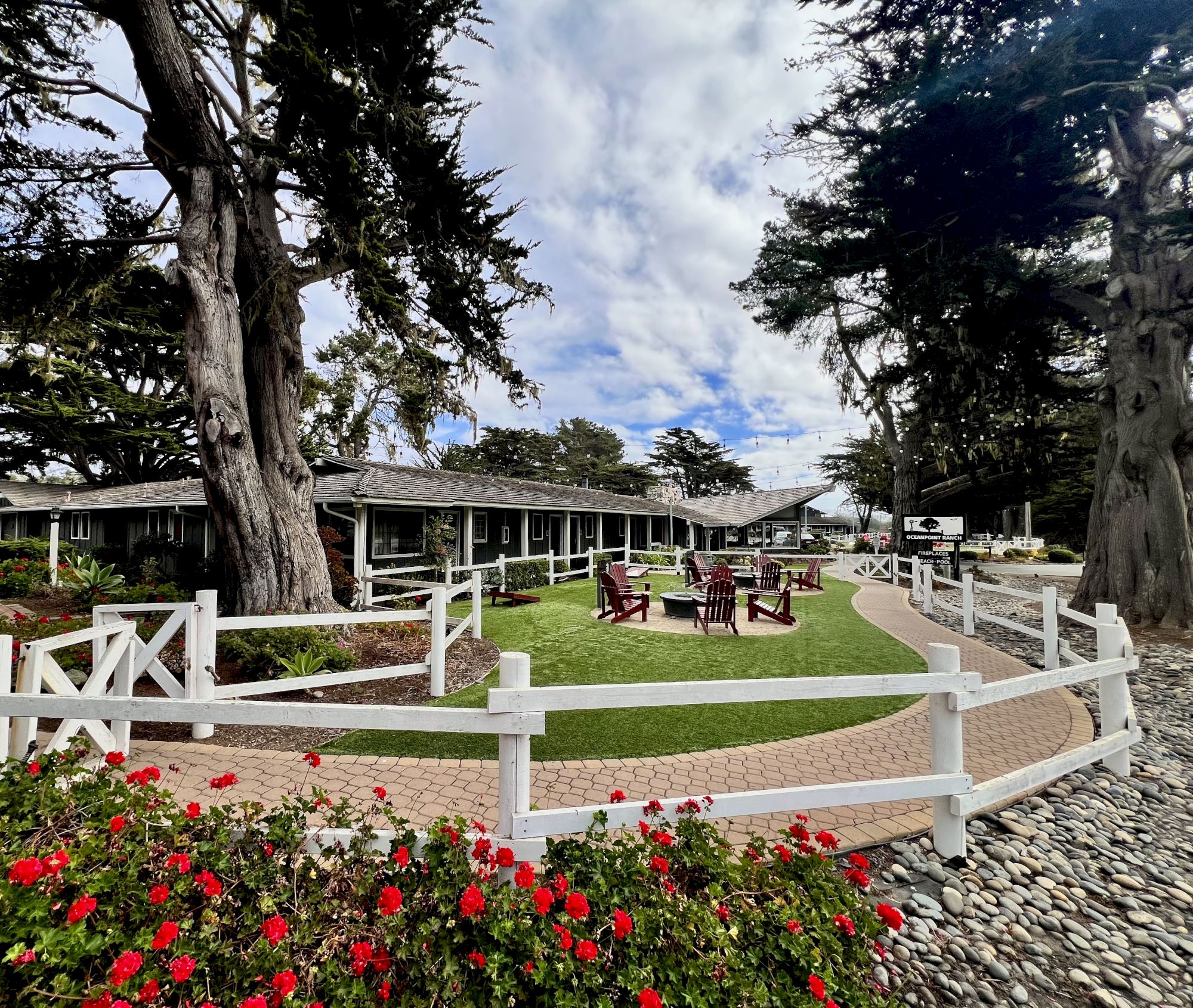 A sunny courtyard with a white split-rail fence, green lawn, outdoor seating, large trees, and colorful flowers surrounding a low stone border.
