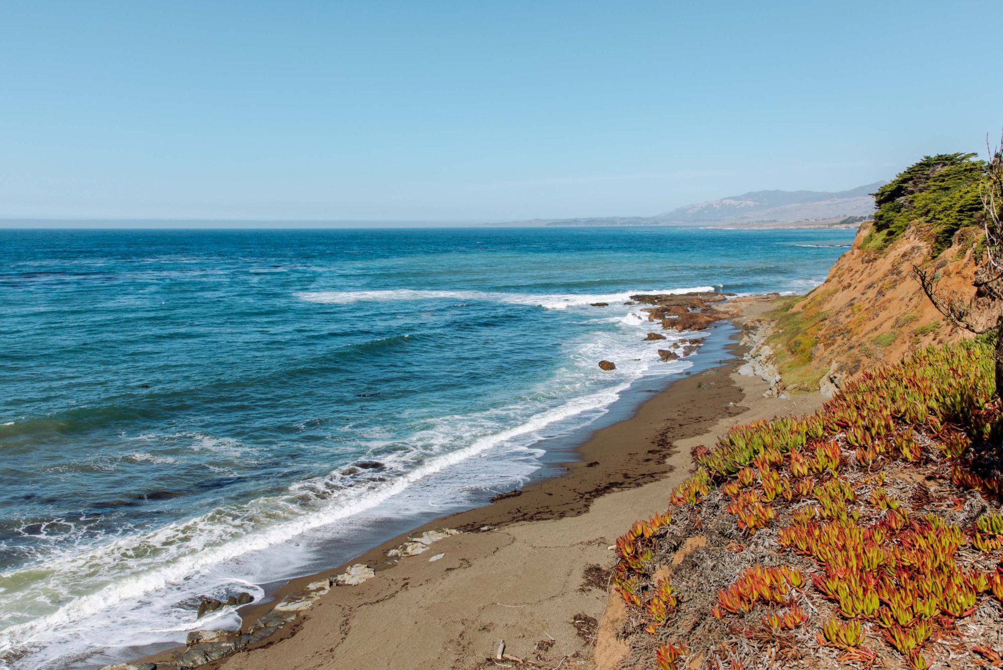 A rocky coastal cliff with orange and green vegetation overlooks a sandy beach and blue ocean waves under a clear sky.