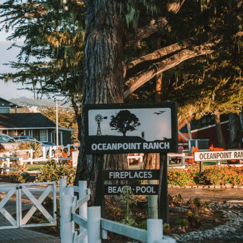 A sign near a tree reads Oceanpoint Ranch, with signs for fireplaces, beach, pool, and a wooden white fence lining a path.