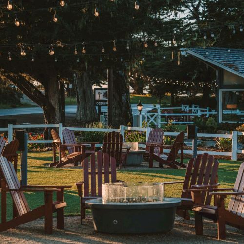 A cozy outdoor fire pit circle with wooden chairs, string lights overhead, trees in the background, and a calm yard vibe evening.