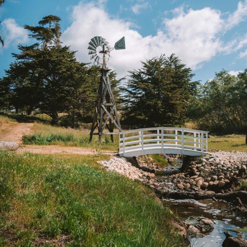 A rustic windmill beside a small white bridge over a rocky stream, with green grass and trees under a bright blue sky.