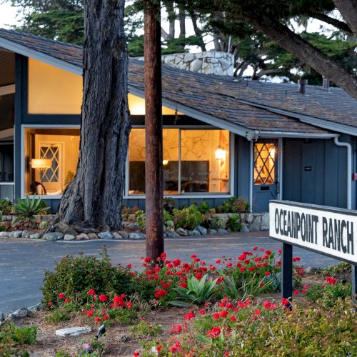 A small, single-story office-style building with a pitched roof, warm interior lights, and a sign reading &ldquo;QUARTPIT RANCH&rdquo; near a garden of red flowers.