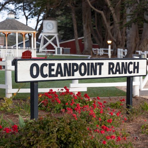 A ranch-style entrance with a white sign reading &ldquo;Oceanpoint Ranch,&rdquo; red chairs, a gazebo, and landscaped flowers near a white fence.