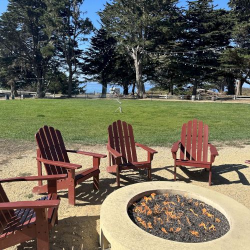 Four red wooden chairs circle a stone fire pit at a sunny park, with green grass, tall trees, and a hint of water in the distance.