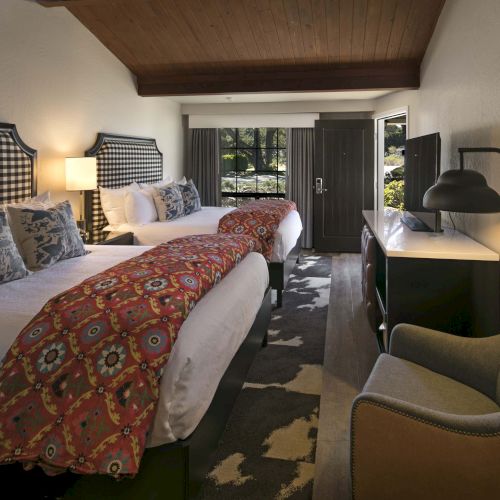A cozy hotel room with two queen beds, patterned headboards, colorful runners, a desk, chair, and a small seating area by the window.