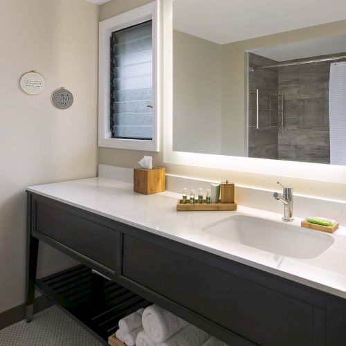 A modern bathroom vanity with a white countertop, dark gray cabinet, large backlit mirror, window, and neatly stacked towels on a lower shelf.