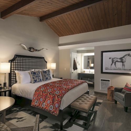 A stylish hotel room with a large bed, patterned bedding, tufted headboard, two lamps, a bench at the foot, and a seating area with a desk and framed horse art, plus warm wood ceiling and ambient lighting.
