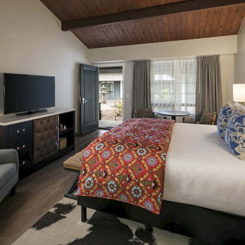 A luxury hotel room with a large bed, patterned bed runner, TV on a dresser, a seating chair, a rug, and a windowed door leading to a balcony.