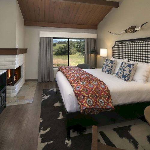 A luxurious hotel bedroom with a tall, checkered headboard, two nightstands, a fireplace, a flat-screen TV, and a large window with curtains overlooking a green lawn, plus a patterned bed runner and decorative pillows.