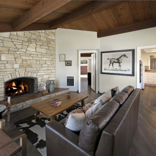 Cozy living room with a stone fireplace, wood beam ceiling, leather sofa, wooden coffee table, and art of a horse; open kitchen in background.