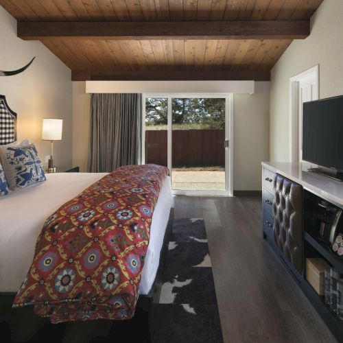 A cozy hotel-like bedroom with a king bed, patterned throw, mounted TV, desk, and a sliding door to a small outdoor area.