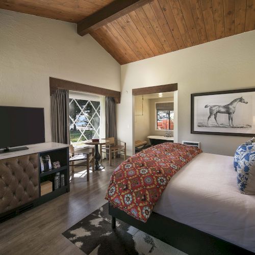 A cozy hotel room with a large bed, patterned red throw, desk, flat-screen TV, and a framed horse picture, under a wooden vaulted ceiling.